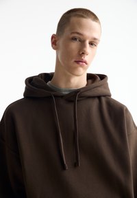 Brauner Hoodie mit großer Fronttasche, Kordelzugkapuze und lockerem Schnitt. Über einem grauen Shirt getragen, das weichen Stoff und ein minimales Design zeigt.