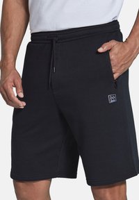 Schwarze Sweatshorts aus weichem Stoff, mit verstellbarem Kordelzug, seitlicher Reißverschluss-Tasche und einem kleinen Logopatch auf der Vorderseite.