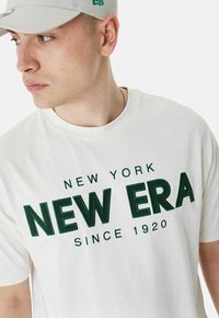 Cremefarbene Baumwoll-T-Shirt mit grün gesticktem Text "NEW YORK NEW ERA SINCE 1920." Rundhalsausschnitt und kurze Ärmel, minimalistisches Design.