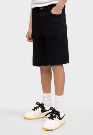 Person iført sorte knælange shorts, hvide crew-sokker og hvide sneakers med sort swoosh og snørebånd, stående med den ene hånd i lommen.