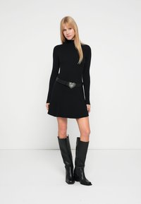 Crna ženska haljina s rebrastim uzorkom i dugim rukavima, proširenim suknjskim dijelom, ukrašena crnim remenom sa kopčom u obliku srca, uparena s crnim čizmama do koljena.