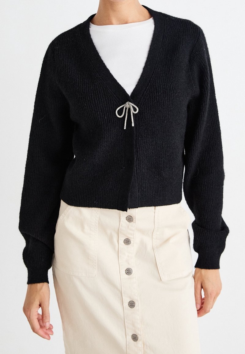 Femme portant un cardigan noir à col en V avec une petite fermeture à nœud papillon par-dessus un haut blanc et une jupe beige taille haute boutonnée.