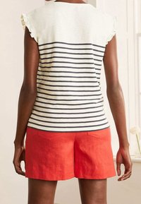 Top sin mangas a rayas en crema y azul marino, con hombros con volantes, combinado con pantalones cortos rojos brillantes que tienen bolsillos traseros y una textura suave.