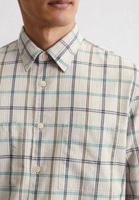 Kurzarm-Hemd mit Knopfverschluss in einem hellen Beige mit kariertem Muster in Blau, Grün und Gelb. Verfügt über einen Kragen und zwei vordere Taschen.