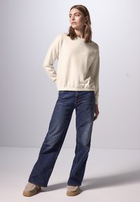 Felpa color crema con orlo arricciato, abbinata a jeans a gamba larga blu. Il modello indossa scarpe slip-on color cammello. Design semplice, stile casual.