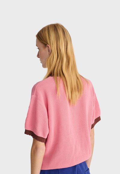 Femme aux longs cheveux blonds portant un pull rose à manches courtes côtelé avec une bande marron, vue de profil sur un fond uni.
