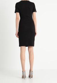 Robe noire ajustée à manches courtes, coupe au genou, détail zippé au dos, portée avec des chaussures à talons gris.