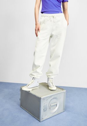 Personne portant un pantalon de survêtement blanc et des baskets montantes blanches, debout sur une boîte métallique grise sur un sol bleu et un mur blanc.