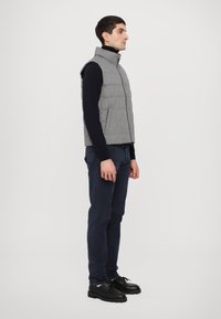 Gilet imbottito grigio con colletto alto, chiusura con zip e senza maniche, indossato sopra una camicia a maniche lunghe scura, abbinato a pantaloni blu navy e scarpe nere.