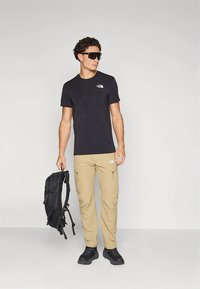 Svart t-skjorte med korte ermer og logo, khakifargede cargobukser med lommer, svarte solbriller og en svart ryggsekk, båret av en mannlig modell.