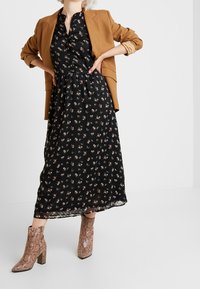 Svart blommig maxiklänning med korta ärmar, insvängd midja och en svävande nederdel, matchad med brun kavaj och stövletter med ormskinnsmönster.