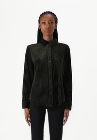Camisa de veludo verde escuro com riscas verticais, fecho frontal com botões, colarinho e bolso. Mangas longas com punhos, textura suave e corte estruturado.