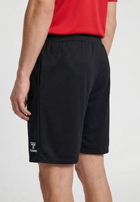 Férfi, fekete Hummel sport rövidnadrágban és piros pólóban, hátulnézetből, egyszerű háttér előtt.