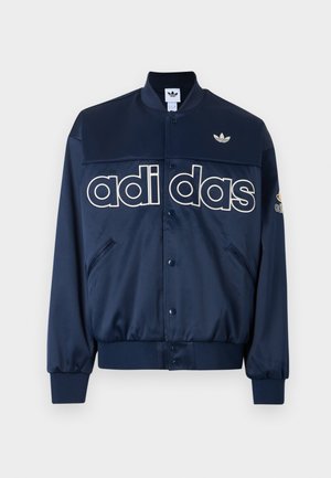 Mand iført navyblå Adidas jakke og bukser med hvidt logo og striber, stående mod en ensfarvet hvid baggrund.