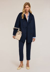 Tummansiniinen tupla-valaistettu takki, jossa on suuret mustat napit, vaaleansinisen paidan päällä, yhdistettynä slim-fit tummansiniisiin housuihin ja beigeen olkalaukkuun.