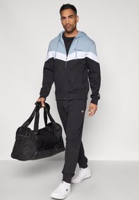 Homem com capuz preto, branco e azul claro, calças combinando e ténis brancos, a carregar uma mochila de ginásio preta contra um fundo simples.