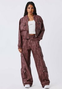 Vrouw met lange vlechten, gekleed in een kastanjebruine cargojack en -broek, met daaronder een witte cropped top en witte sneakers, staande tegen een effen achtergrond.