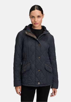 Frau trägt eine marineblaue gesteppte Jacke mit Kapuze, vorderen Knöpfen und Klappentaschen, steht vor einem schlichten hellen Hintergrund.