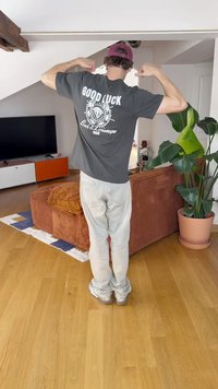 Grå t-skjorte med hvit grafisk tekst "GOOD LUCK" og emblem på baksiden, sammen med beige bukser og sølvfargede sko, i et hjemmemiljø.