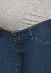 Blå denimjeans med en strukturerad yta, med knappstängning och gula sömmar längs midjan och fickorna. Grått tyg synligt ovanför.