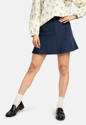 Marineblauwe uitlopende minirok met een gladde textuur, gedragen met een lichtgele blouse met patroon en zwarte loafers, gecombineerd met witte geribbelde sokken.