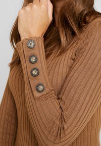 Maglietta a maniche lunghe marrone a coste con quattro bottoni decorativi sul polsino, evidenziando il tessuto texture e il design strutturato.