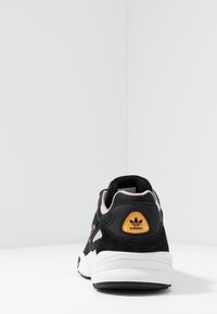 Svart mockaskor med flerfärgade accenter, med en vit mellansula och en guldbrodyr vid hälen. Texturerat nät på sidorna.