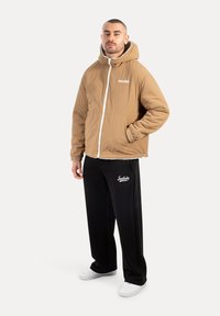 Chaqueta acolchada marrón con capucha, cremallera blanca y logo bordado, combinada con pantalones de chándal negros que llevan el mismo logo y zapatillas blancas.