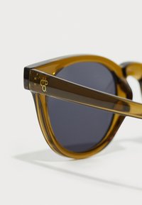 Sunglasses with amber translucent plastic frame, round dark lenses, and gold logo detail on the arm. Smooth texture with a modern design.