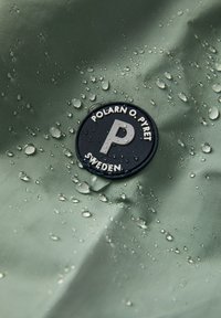 Vihreä vedenpitävä kangas, jossa on vesipisaroita; mukana pyöreä musta logo, jossa lukee "POLARN O. PYRET" ja erinomainen "P" -muotoilu.