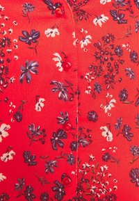 Rød bluse med blomster, der har små lilla og hvide blomster, fremstillet i et struktureret stof med røde knapper, der løber ned ad forsiden.
