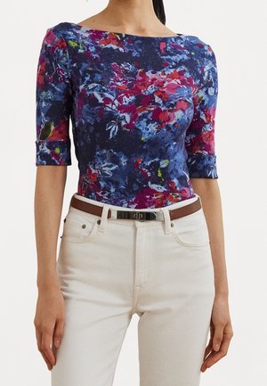 Kobieta ubrana w niebieską kwiecistą bluzkę z półrękawami, włożoną do białych spodni z brązowym paskiem, stojąca z rękami swobodnie opuszczonymi wzdłuż ciała.