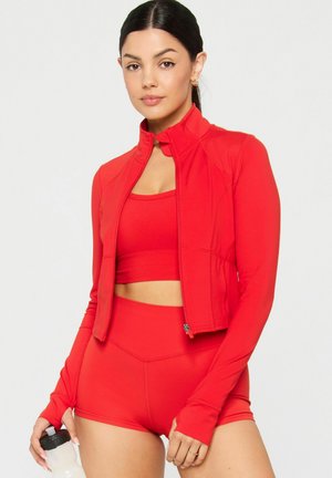 Woman wearing red athletic crop top, zip-up jacket, and shorts, holding a white water bottle with black lid, standing against white background.