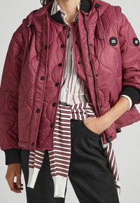 Quilted burgunderfarbener Jacke mit schwarzen Akzenten, Druckknöpfen, Kapuze und gemusterter Fütterung; getragen über einem gestreiften Hemd und schwarzen Hosen.