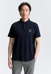 Marineblauwe polo met een kraag, drie-knopen sluiting en een klein bedrukt logo. Korte mouwen en een fijne katoenen textuur.
