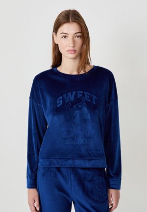 Junge Frau in einem marineblauen Samt-Sweatshirt und passenden Hosen mit geprägtem Teddy-Bär und dem Text "SWEET dreams".