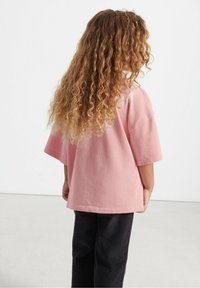 Lyserød bomulds t-shirt med en afslappet pasform og korte ærmer, parret med teksturerede sorte fløjl bukser. Udsigten er fra bagsiden.