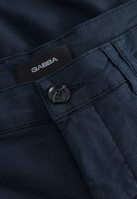 Tamsiai mėlynos tekstūruotos kelnės su juodu sagos užsegimu ir matoma juoda etiketė su užrašu „GABBA“ diržo viduje.