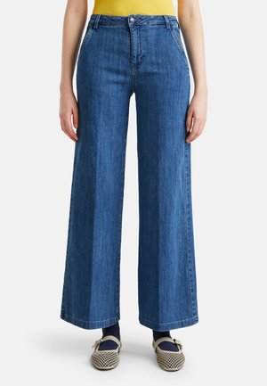 Person trägt hoch taillierte, weit geschnittene blaue Jeans, gelbes Oberteil, schwarze Socken und beige geflochtene Mary Jane Schuhe.