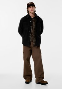 Sort fleece-skjortejakke med to brystlommer over en brun ternet skjorte, kombineret med brune cargobukser og brune støvler.