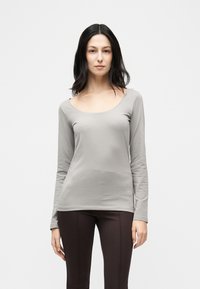 Γυναίκα με ίσια σκούρα μαλλιά που φοράει ανοιχτό γκρι μακρυμάνικη μπλούζα με στρογγυλή λαιμόκοψη και σκούρα εφαρμοστά παντελόνια σε απλό λευκό φόντο.