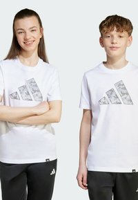 White cotton t-shirts feature a grey camo pattern in the shape of three stripes on the front. Paired with black pants.