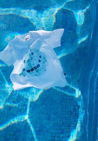 Camiseta blanca de algodón con un patrón de mosaico en azul y turquesa, flotando en agua clara, con ondas que reflejan la luz.