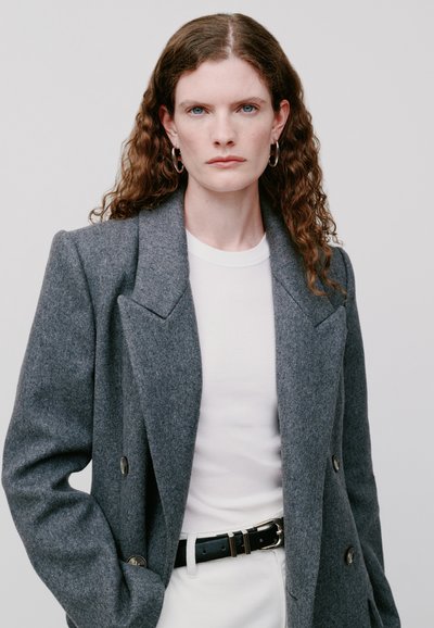 Blazer grigio in lana con design doppiopetto, revers a punta e bottoni neri, abbinato a una camicia bianca e una cintura nera per un contrasto.