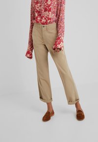 Beige bomullbyxor med uppvikta cuffar, kombinerade med rostfärgade mocka-loafers. Modellen har på sig en röd blommig skjorta med lång ärm.