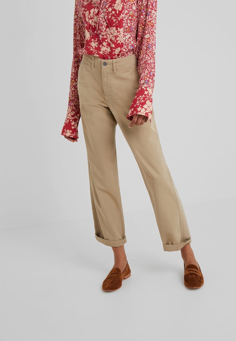 Beige bomullbyxor med uppvikta cuffar, kombinerade med rostfärgade mocka-loafers. Modellen har på sig en röd blommig skjorta med lång ärm.