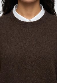 Gros plan d'une personne portant une chemise blanche à col sous un pull en tricot brun foncé, montrant le cou et le haut de la poitrine.