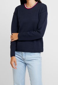 Pull-over bleu marine avec un col rond orné d'une bordure rayée tricolore. Confectionné en tissu texturé, manches longues et coupe décontractée.