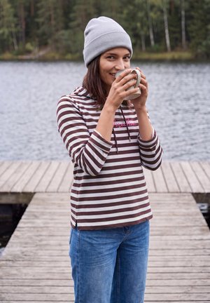 Femme portant un bonnet gris et un sweat à capuche rayé boit dans une tasse en se tenant sur un ponton en bois au bord d’un lac avec des arbres en arrière-plan.