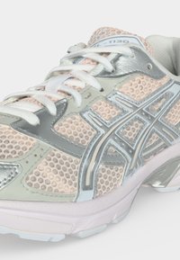 Jasnoróżowy i srebrny sportowy sneaker z białymi sznurowadłami i siatkową cholewką, z logo marki po boku i numerem modelu na języku.